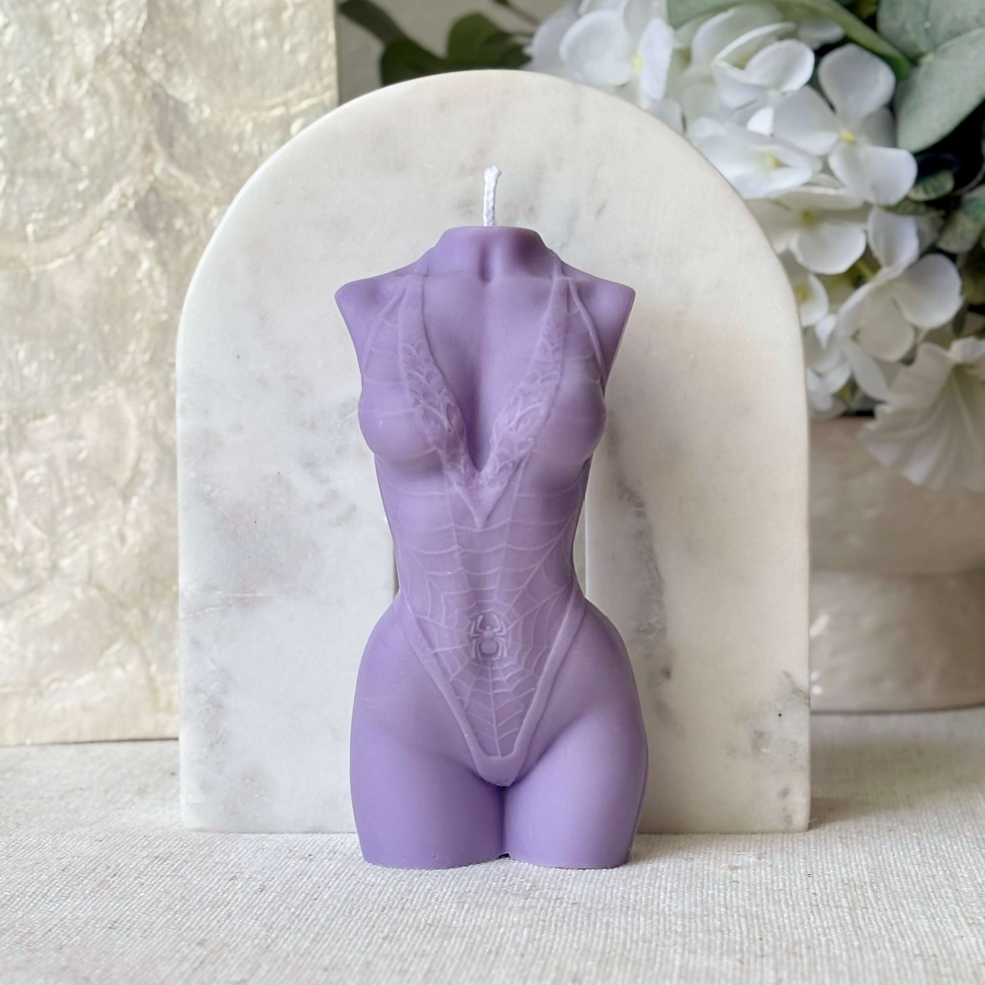 Purple female torso candle handmade from soy wax, artistic body sculpture accent with white flowers in background.