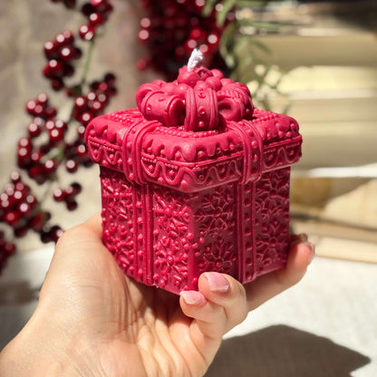 Red handmade candle shaped like a gift box, held in hand with red berries in background.