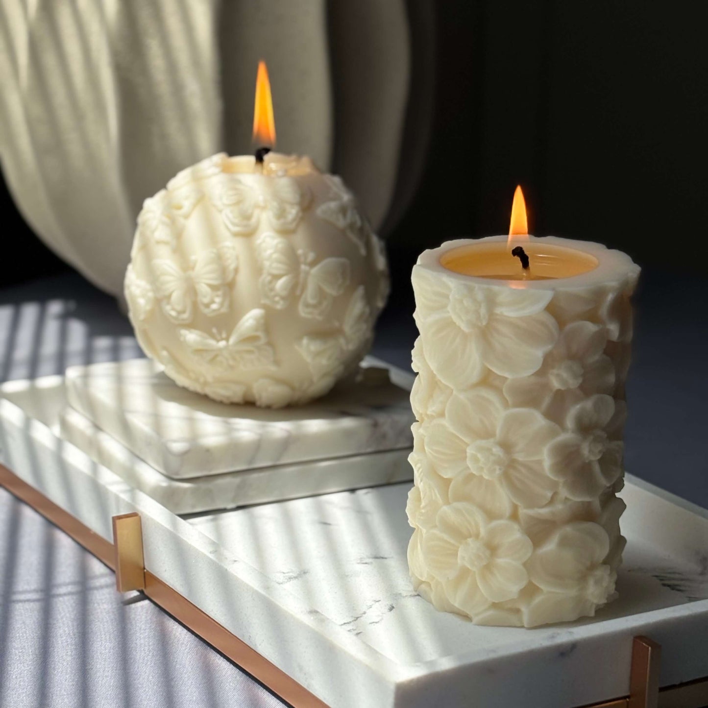 Pair of sculpted soy candles – floral pillar and butterfly sphere burning on tray	
