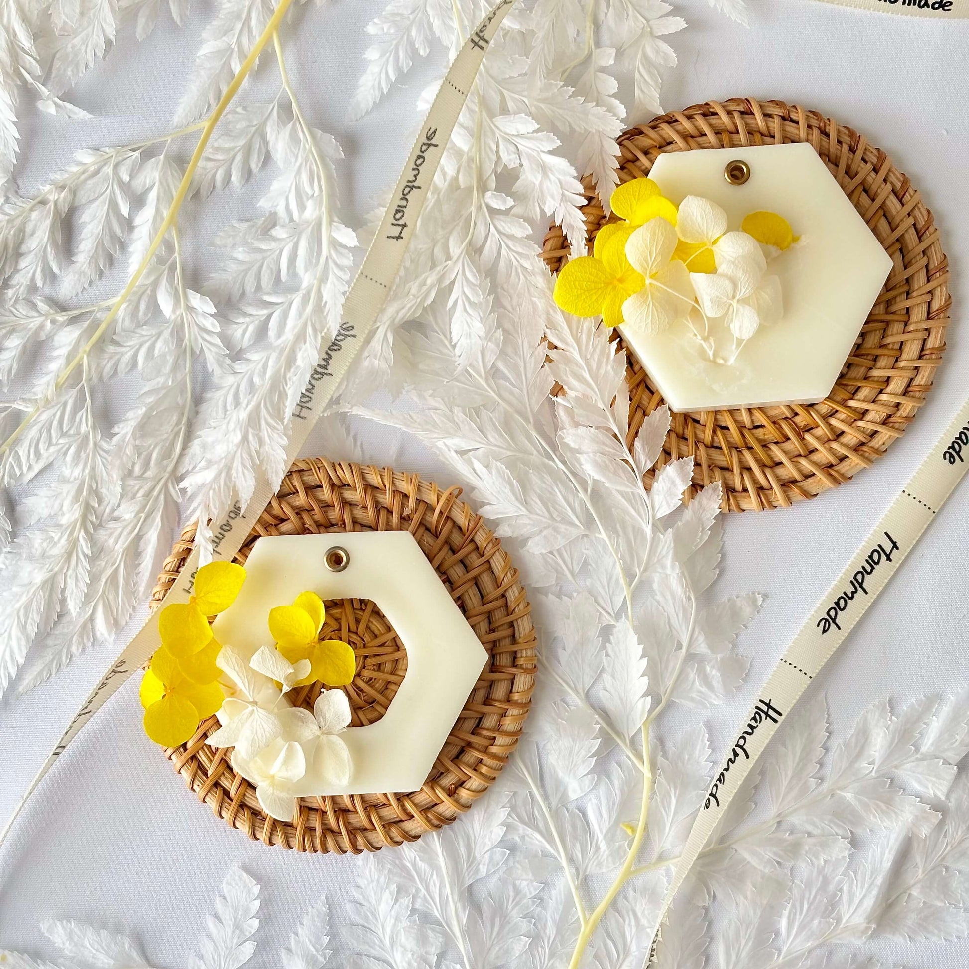 Flat lay of two handmade botanical soy wax sachets with dried yellow and white flowers, styled on woven rattan coasters with a handmade ribbon accent.