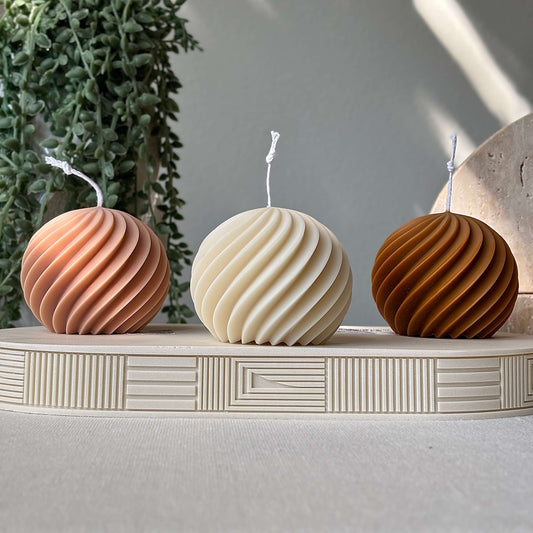 Three spiral soy wax candles in peach, beige and honey tones on a decorative stand with greenery in the background.