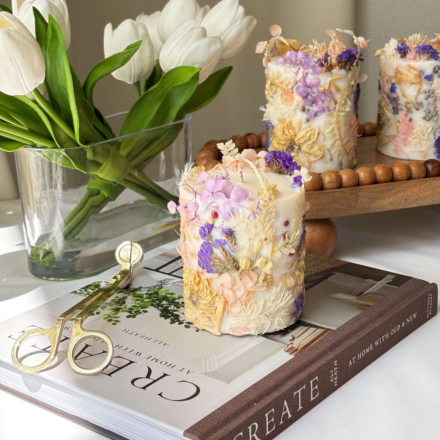 Three handmade botanical soy candles with dried pastel flowers in purple, pink, and cream tones, styled on books with tulips and gold wick trimmer.