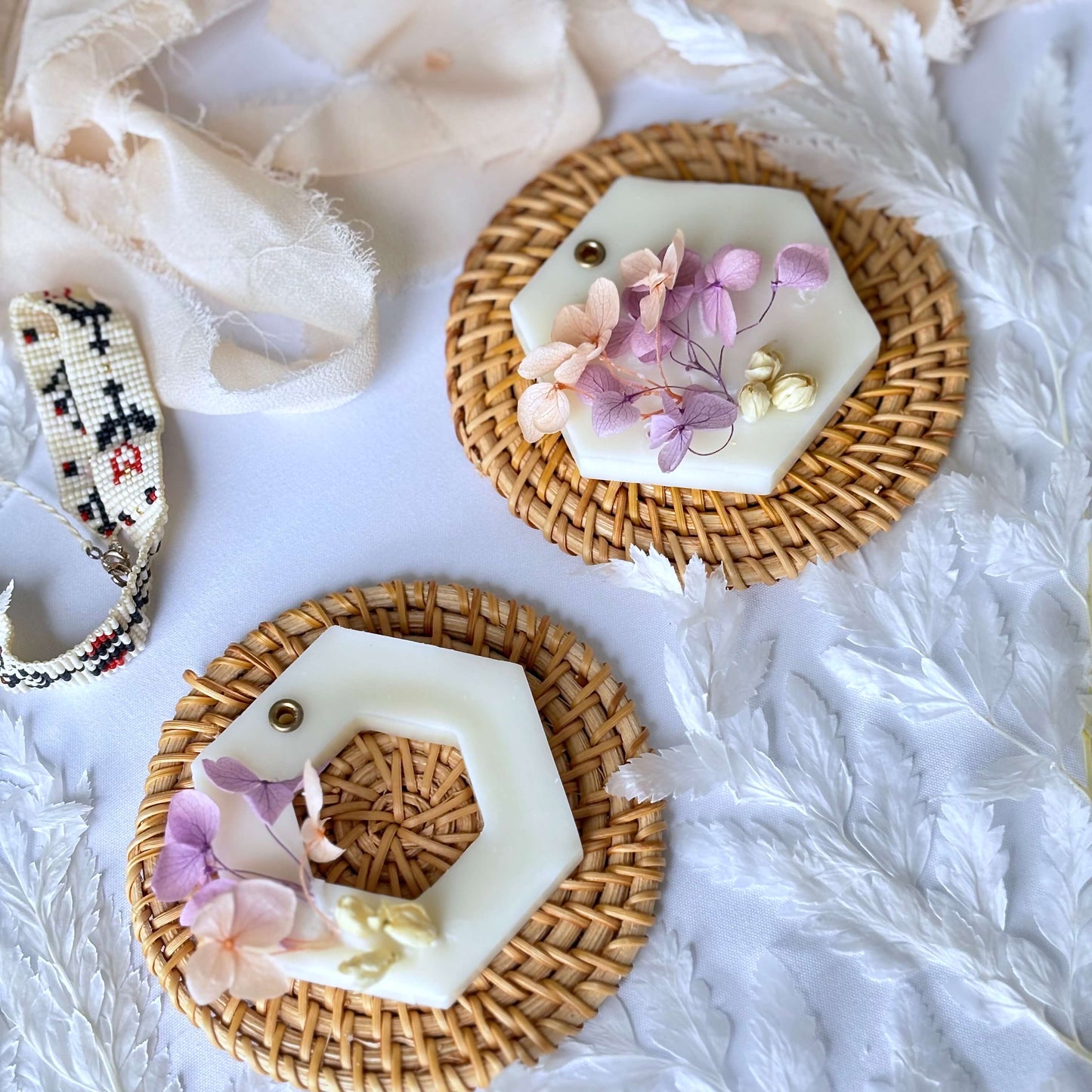 Two handmade soy wax sachets with dried pink and lavender flowers, placed on rattan mats and styled with soft fabric and white leaves for a cozy flatlay.