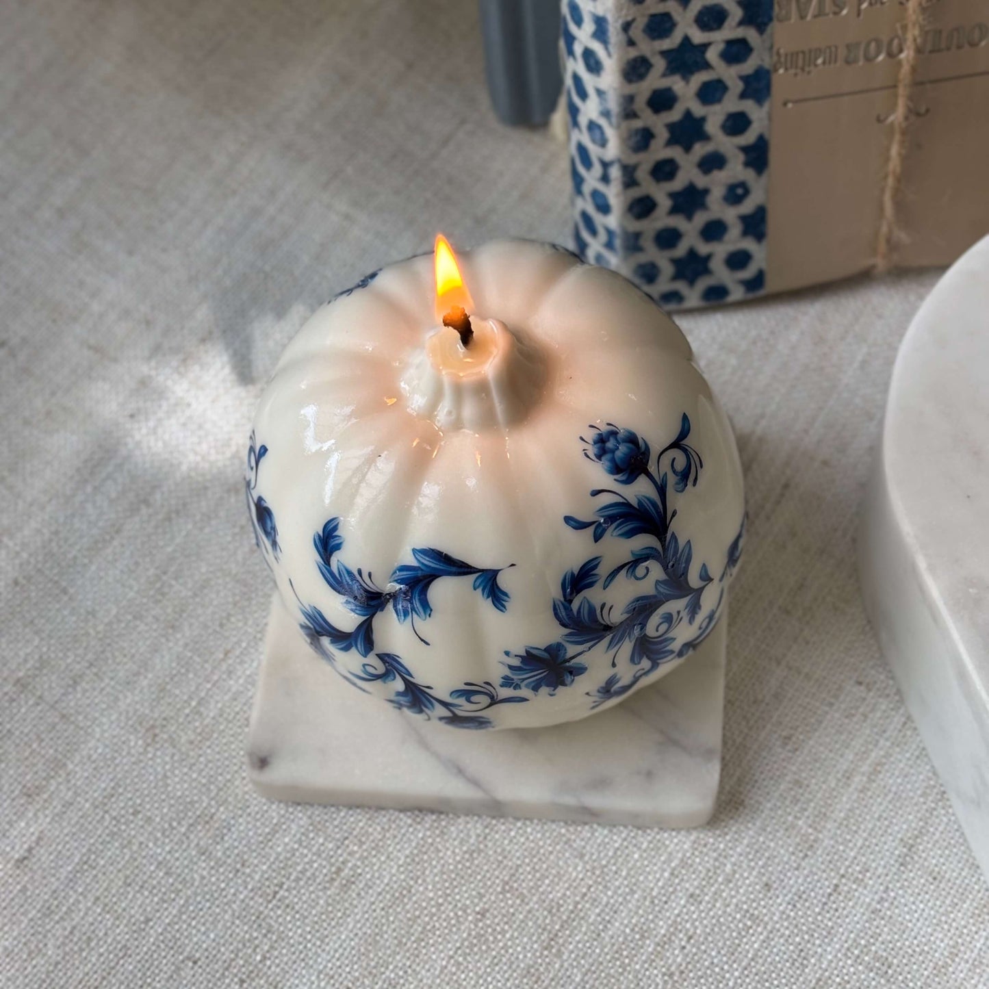 Unscented round toile-style candle with a lit wick and blue floral accents, displayed on a stone tray for neutral interior styling.