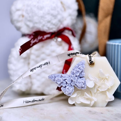 Close-up of handmade floral wax sachet with lavender butterfly next to plush teddy towel and ceramic mug — a charming touch in a premium self-care gift box for her